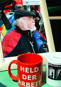Stillleben. Vor einem Bild Manni Demmers steht eine rote Tasse mit der Aufschrift »Held der Arbeit«. Auf der Spendendose daneben kann man »Kultur A...« erkennen.