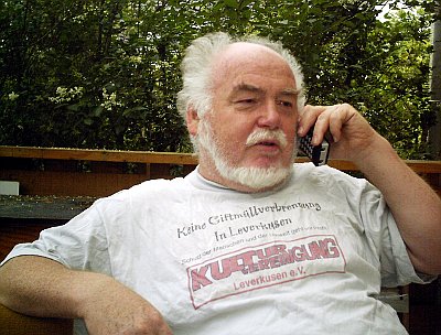 Manni Demmer telefoniert. Auf dem weißen T-Shirt mit dem Logo der Kulturvereinigung stehen die Forderungen »Keine Giftmüllverbrennung in Leverkusen« und »Schutz der Menschen und der Umwelt geht vor Profit«.
